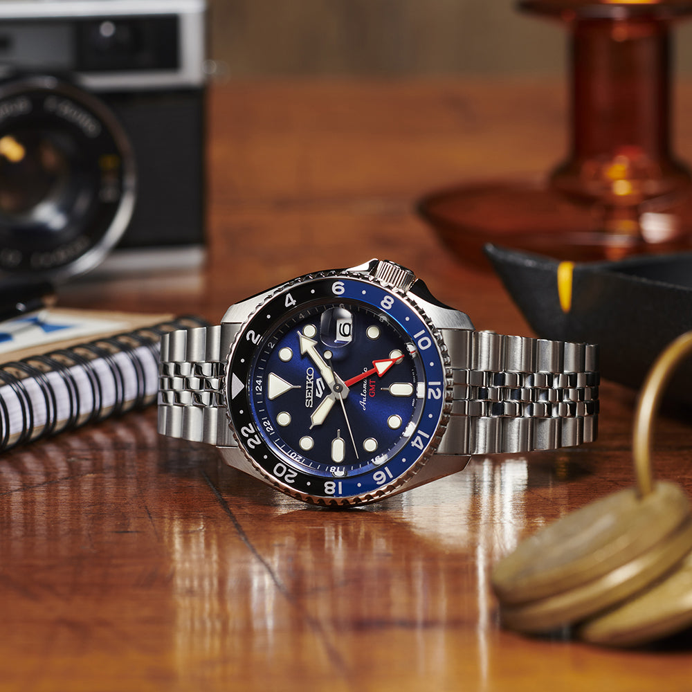 Een Seiko 5 Sports herenhorloge SSK003K1 met een zilveren metalen band en blauwe wijzerplaat rust op een houten tafel. Het beschikt over een waterdichtheid van 10ATM, een datumweergave en lichtgevende markeringen. Op de achtergrond staan een vintage camera, notitieboekje en een klein metalen voorwerp.