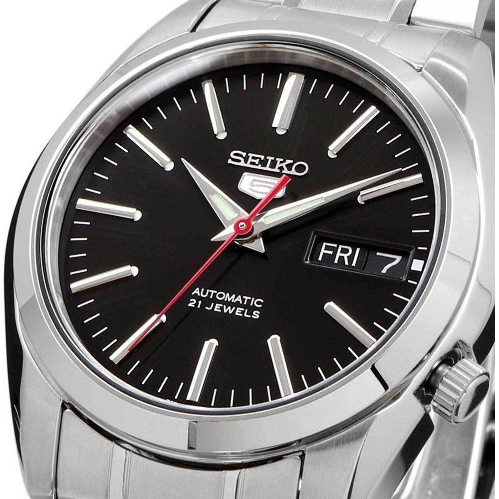 Een close-up van het Seiko heren horloge SNKL45K1 toont zijn roestvrijstalen kast. De zwarte wijzerplaat heeft zilveren uurmarkeringen, een rode secondewijzer en een dag-datumvenster met FRI 7. Dit stijlvolle horloge is trots gelabeld als Automatic 21 Jewels.