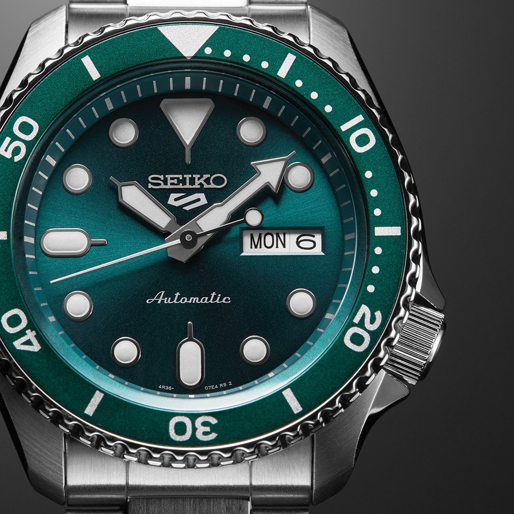 Close-up van de Seiko 5 Sports SRPD61K1, een duikhorloge met een groene wijzerplaat. Het heeft zilveren uurmarkeringen, een dag-datumweergave met MON 6, een draaiende lunette met cijfers en een roestvrijstalen band naast de getextureerde kroon.