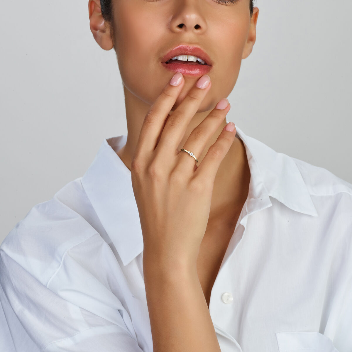 Een vrouw in een wit overhemd tikt zachtjes over haar gezicht en toont de Van der Kooij Collectie ring trilogie zirkonia 14K geelgoud aan haar vinger. Haar lippen zijn lichtjes gespleten en haar nagels zijn netjes gemanicuurd.