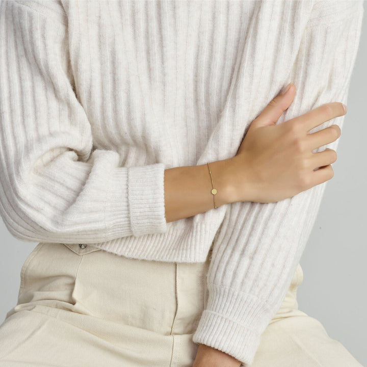 Een persoon in een crèmekleurige geribbelde trui en lichte broek zit met één arm gekruist en draagt de Van der Kooij Collectie armband zirkonia 16,5 - 17,5 - 18,5 cm 14K geelgoud met een kleine ronde zirkonia bedel.