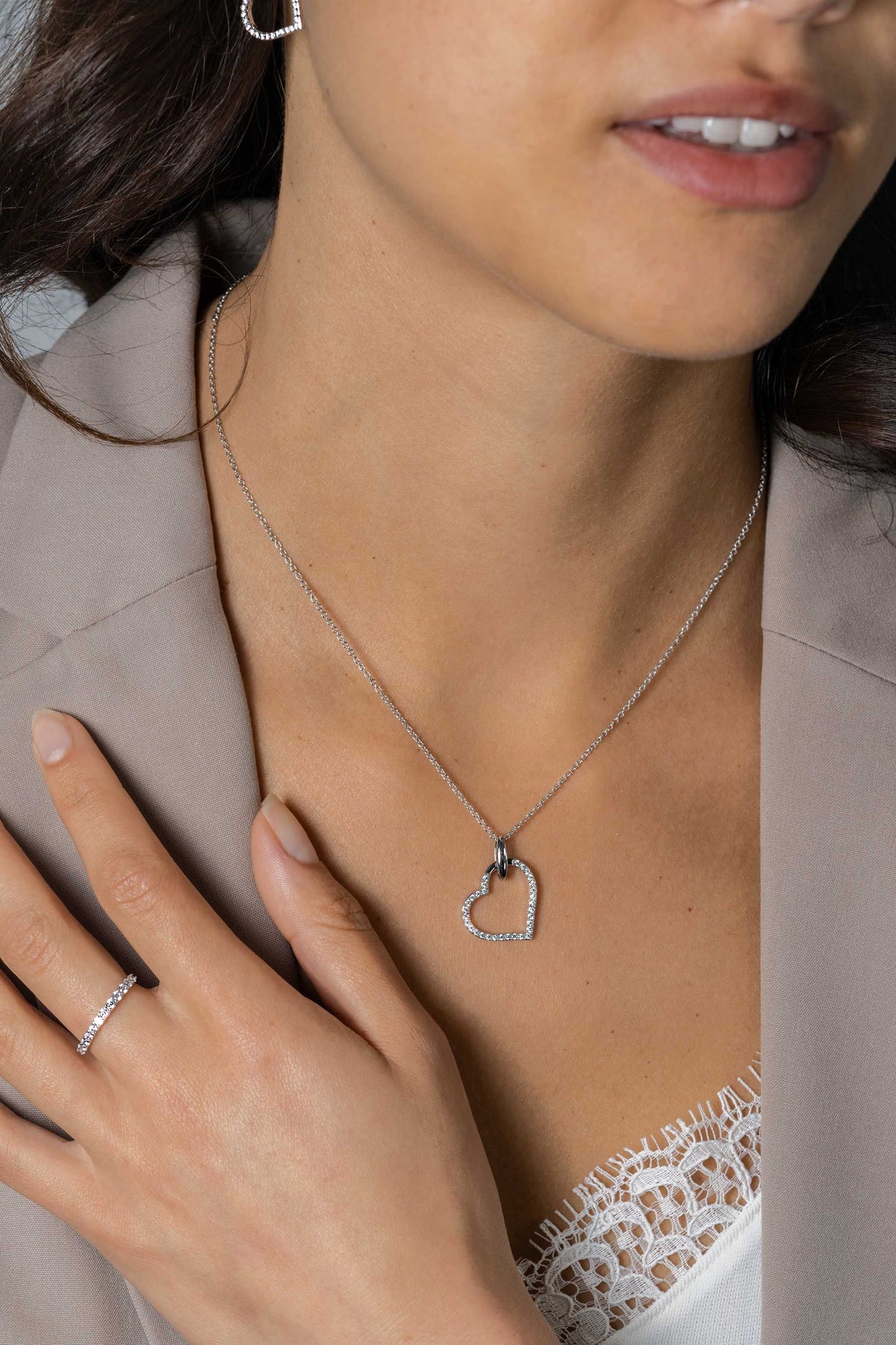 Een vrouw in een beige blazer en wit kanten topje showt de Zinzi Hangers Zilver ZIH2447 in een luxe hartontwerp, gecombineerd met een bijpassende zilveren ring versierd met witte zirkonia's, haar hand zachtjes rustend op haar borst.