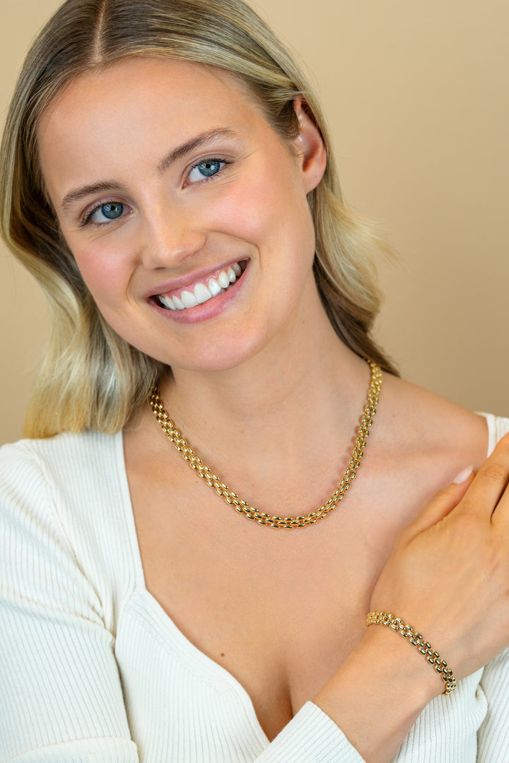 Een blonde vrouw met blauwe ogen in een wit topje lacht naar de camera, draagt een Zinzi Ketting Gold Plated ZIC2764G en bijpassende armband van Zinzi, haar linkerhand raakt zachtjes haar rechterschouder aan tegen een beige achtergrond.