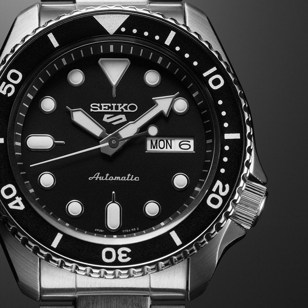 Close-up van een Seiko 5 Sports SRPD55K1 horloge, met een zwarte wijzerplaat met opvallende witte uurmarkeringen, dag-datumweergave en minuten-gemarkeerde roterende lunette. De zilveren roestvrijstalen kast en armband versterken het ontwerp.