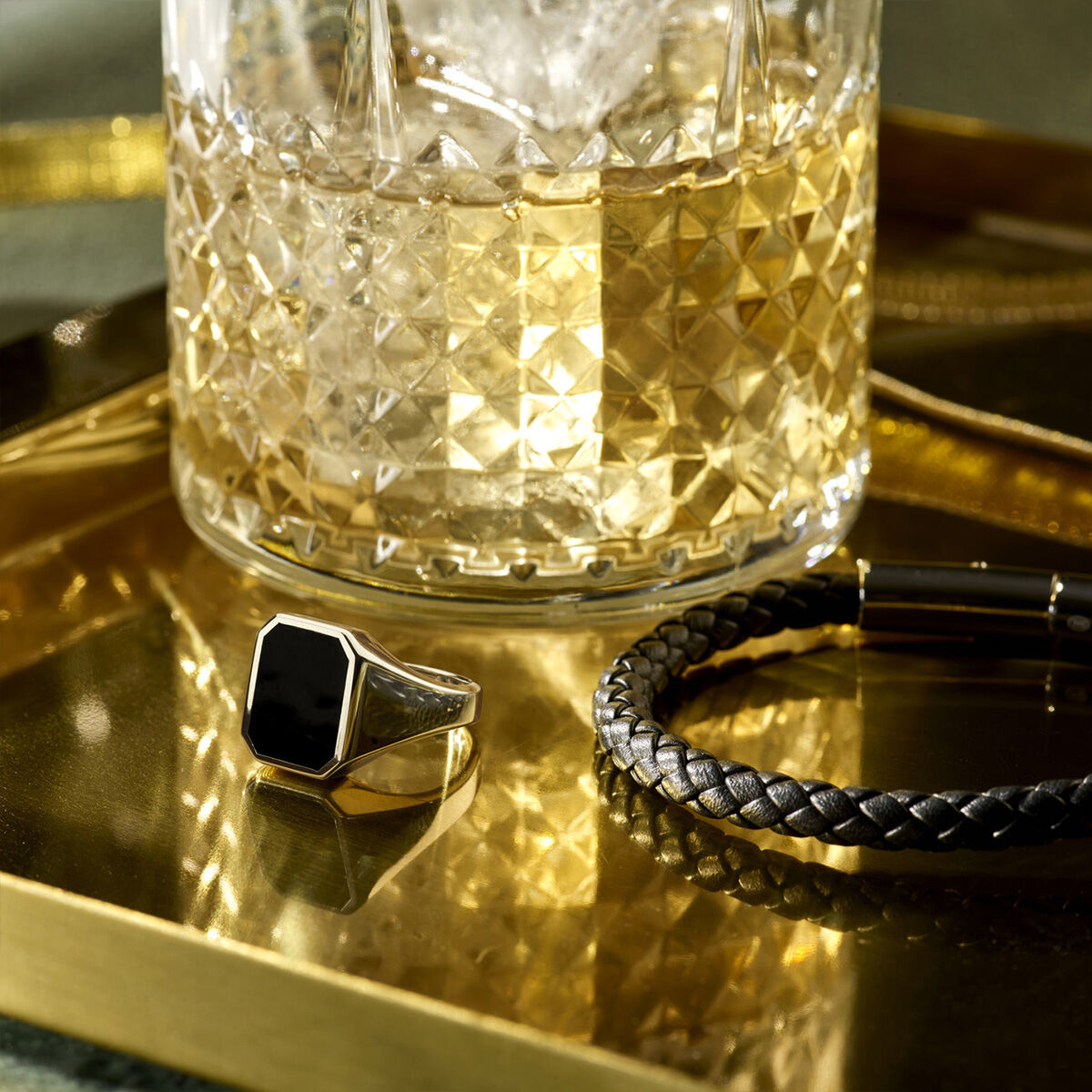 Een gefacetteerd glas whisky met ijs staat op een gouden dienblad naast een Van der Kooij Collectie zegelring massief onyx 13 x 11mm zilver gerhodineerd en een zwarte gevlochten armband met metalen sluiting.