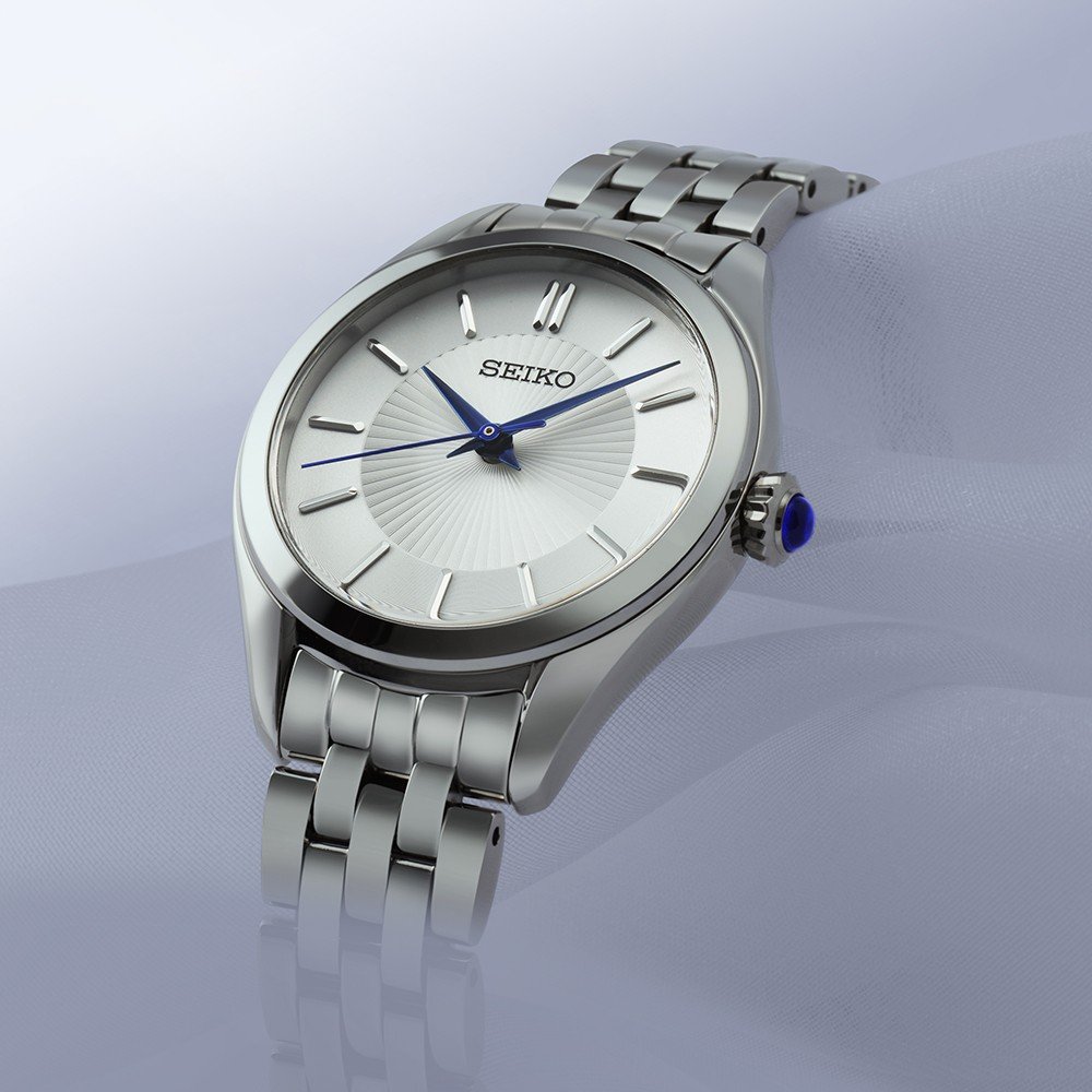Het Seiko heren horloge SUR557P1 heeft een witte wijzerplaat met zilveren markeringen en opvallende blauwe wijzers. Seiko siert de wijzerplaat, aangevuld met een roestvrijstalen band. Het beschikt over 10ATM waterbestendigheid en krasbestendig saffierglas, afgezet tegen een zachte wazige gradiëntachtergrond.