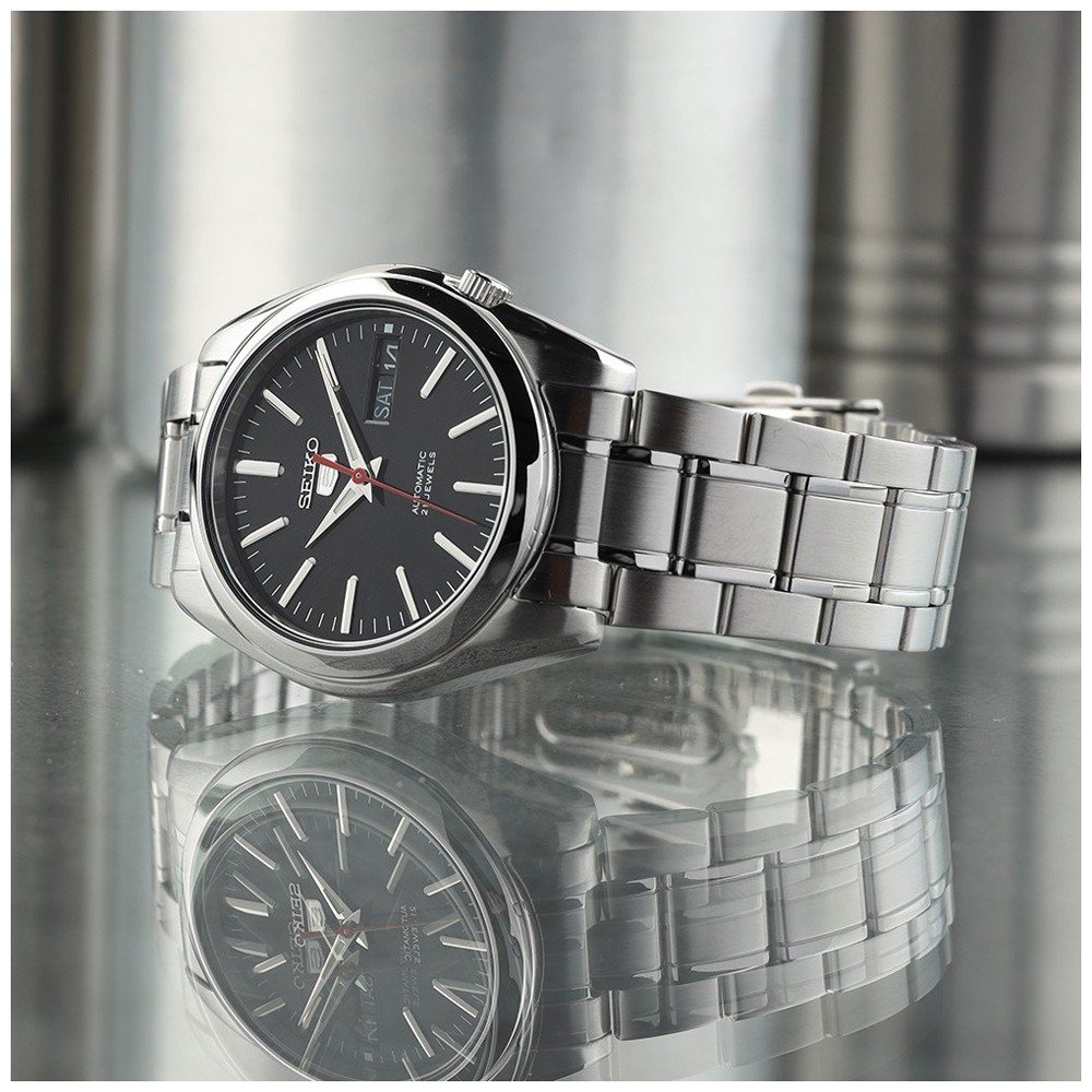 Het Seiko heren horloge SNKL45K1, met een zwarte wijzerplaat, zilveren uurmarkeringen, roestvrijstalen band, automatisch uurwerk en dag-datumaanduiding, wordt afgebeeld op een reflecterend oppervlak.
