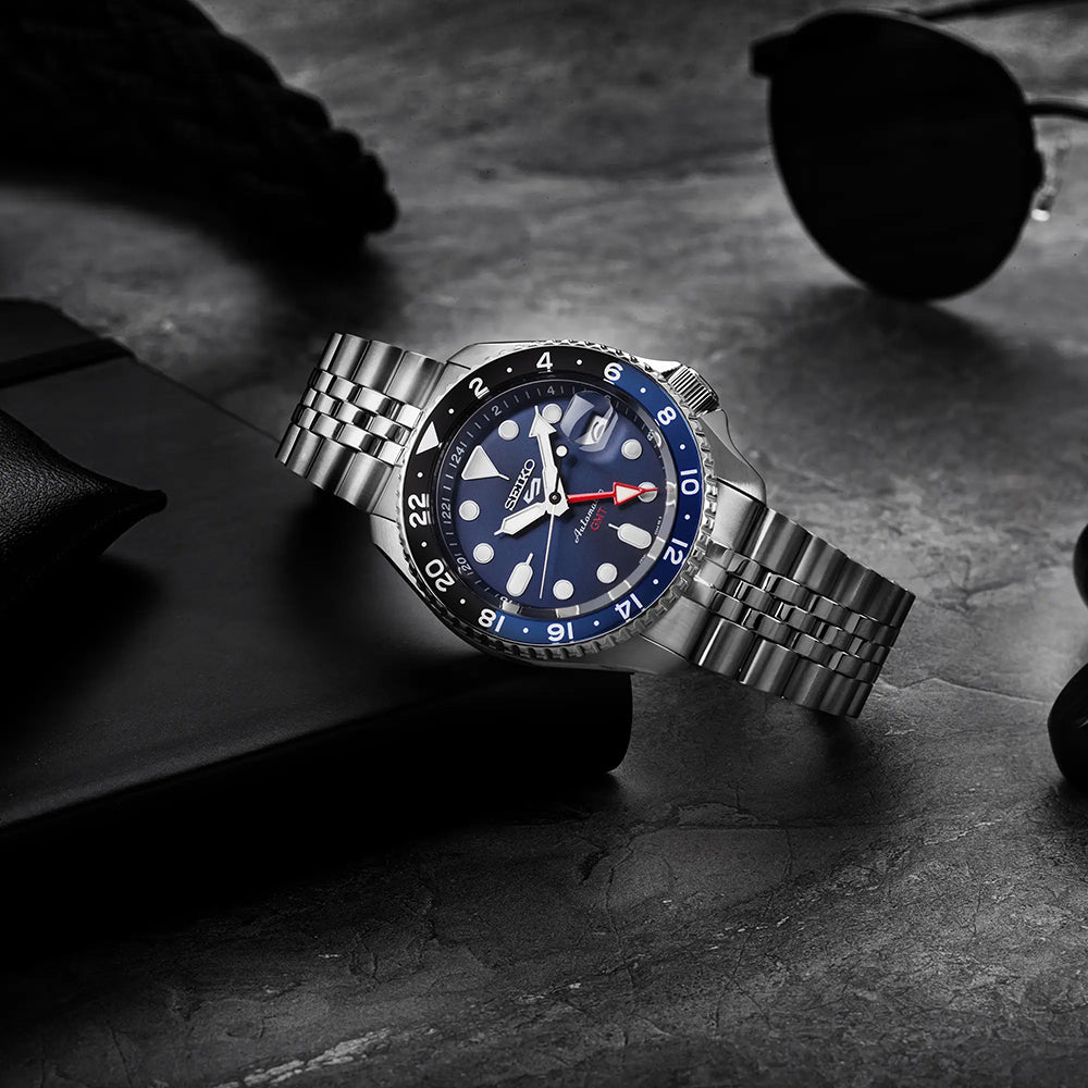 Op een donker oppervlak toont het Seiko 5 Sports herenhorloge SSK003K1, met een automatisch uurwerk en blauw-rode lunette, zijn blauwe wijzerplaat en zilveren metalen band naast een zwarte zonnebril en een notitieboekje. Deze strakke opstelling is compleet met een waterdichtheid van 10ATM.