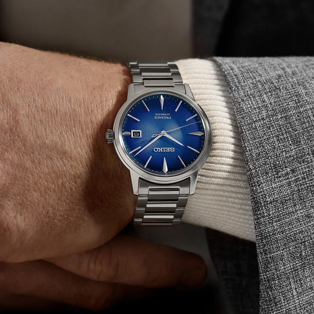 Een persoon in een grijs pak draagt een Seiko Presage heren horloge SRPJ13J1 met een roestvrijstalen kast, blauwe wijzerplaat, datum op 3 uur, zilveren markeringen, drie wijzers en merk-/modelinformatie op de wijzerplaat.
