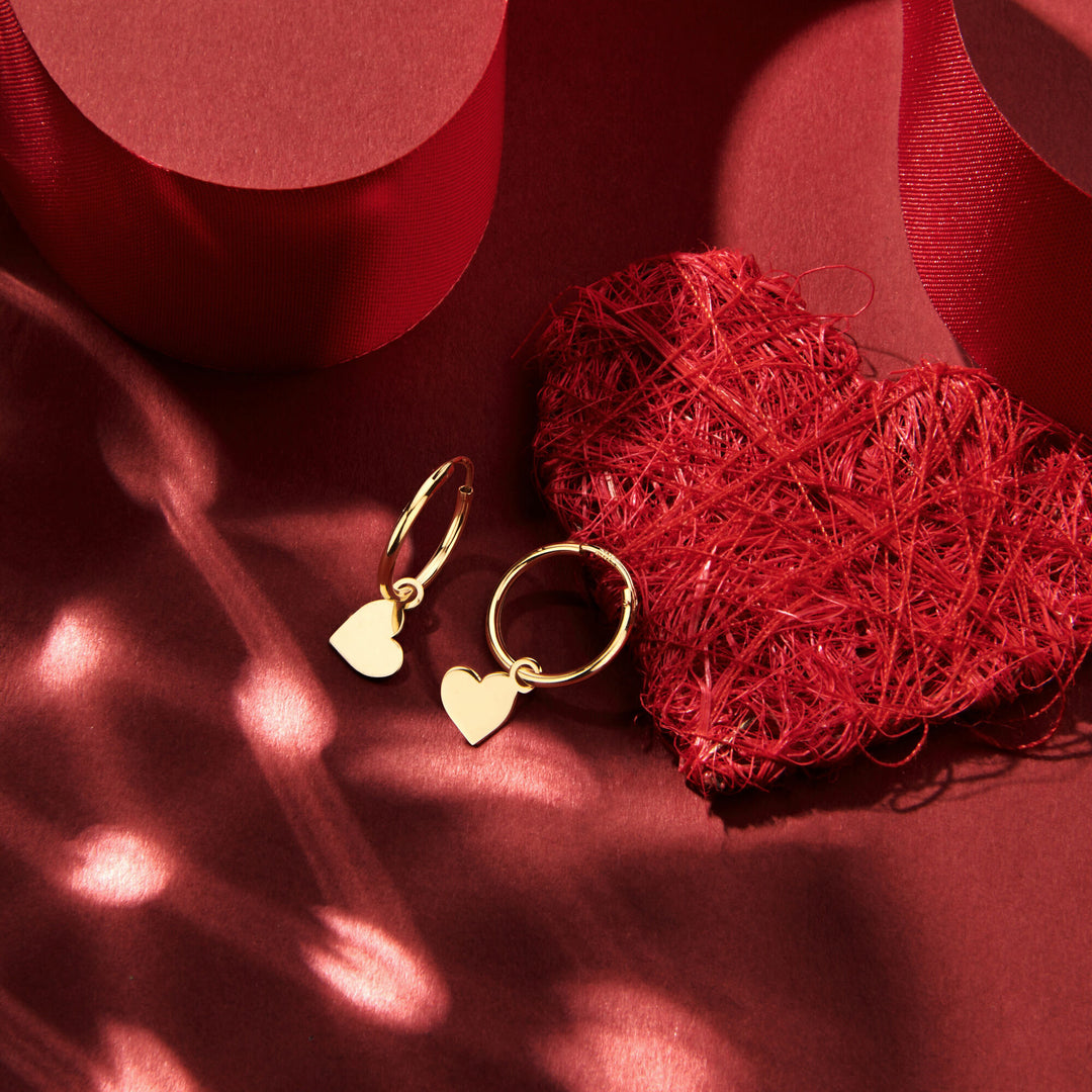 Van der Kooij Collectie oorringen hart 14K geelgoud met delicate hart bedels liggen bij een gestructureerd rood hart en lint in zacht zonlicht - perfect voor Valentijn en vakkundig handgemaakt.