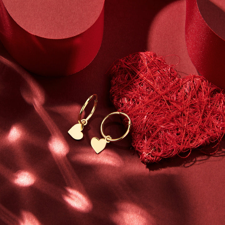 Van der Kooij Collectie oorringen hart 14K geelgoud met delicate hart bedels liggen bij een gestructureerd rood hart en lint in zacht zonlicht - perfect voor Valentijn en vakkundig handgemaakt.