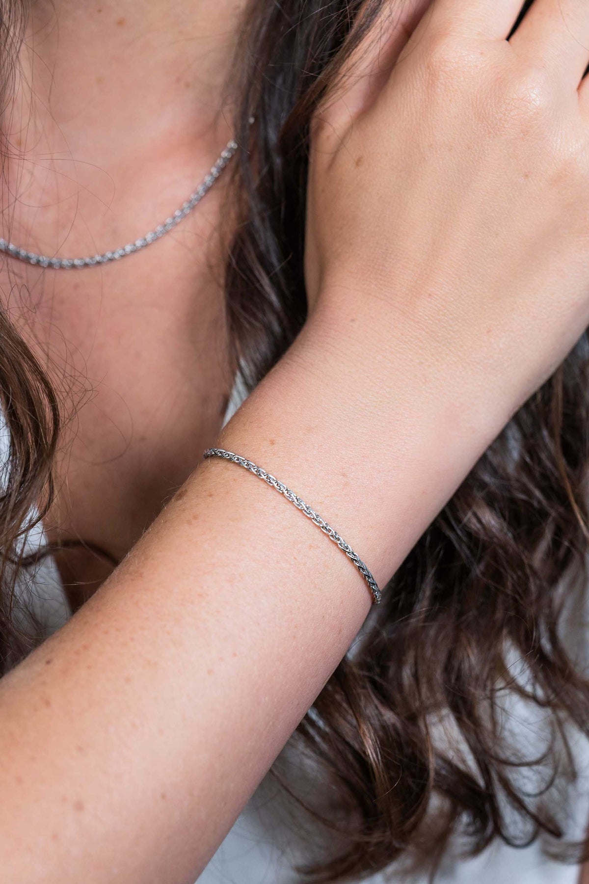 Een persoon met lang bruin haar draagt een elegante zilverkleurige armband vergelijkbaar met de Zinzi Armband Zilver Zia1931 en een bijpassende ketting. Hun hand is bij hun nek, gekleed in een wit topje.