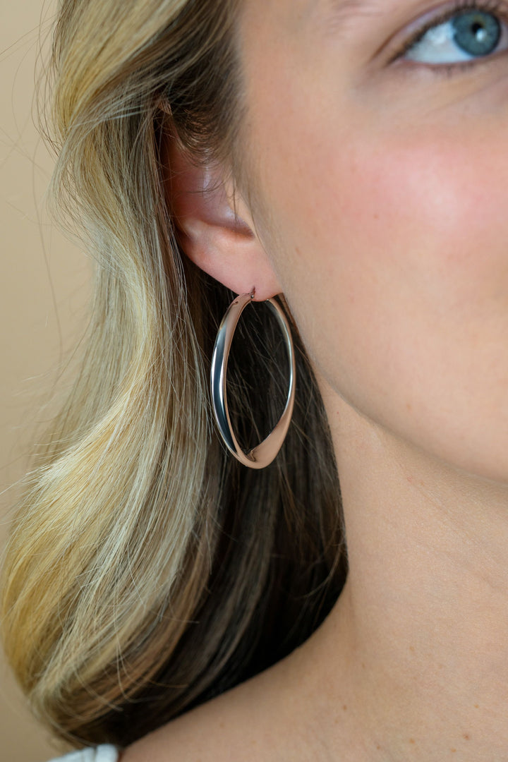 Close-up van een vrouwenoor met lang blond haar, dat grote, glanzende Zinzi ZINZI zilveren oorringen ZIO2702 draagt. Slechts een deel van haar gezicht is zichtbaar, met de nadruk op de vergulde oorbellen en de onderste helft van haar oog.