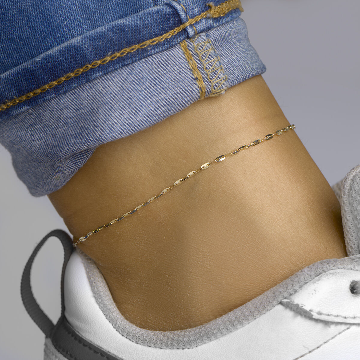 Een close-up van een enkel met het Van der Kooij Collectie enkelbandje plaatjes 1,5 mm 24-26 cm 14K geelgoud, gestyled met een blauwe jeans met gele stiksels en een witte sneaker met grijze accenten.