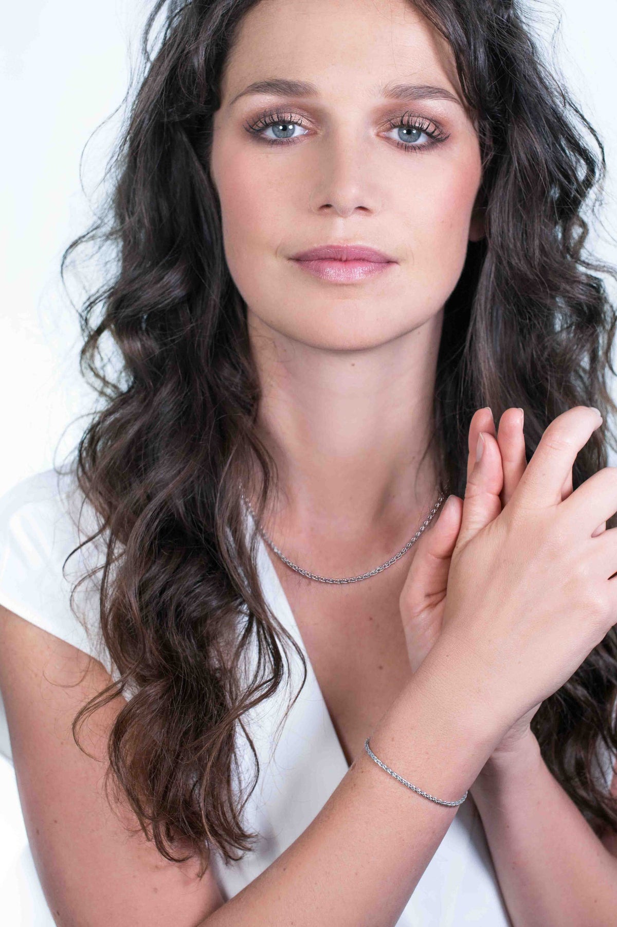Een vrouw met lang, krullend bruin haar, draagt een wit topje en een delicate zilverkleurige ketting. Ze poseert tegen een zachte, wazige witte achtergrond, haar handen lichtjes gevouwen voor haar. Om haar pols draagt ze de Zinzi Armband Zilver Zia1931 van Zinzi, gecombineerd met een subtiele glimlach.