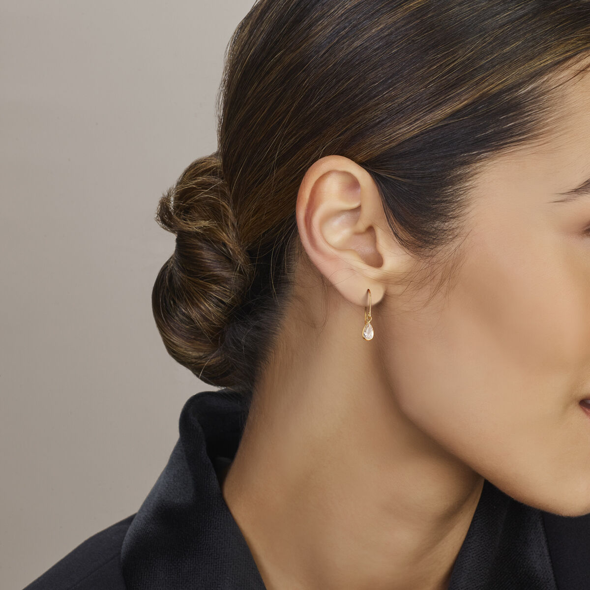 Close-up van een vrouw met donker haar in een lage knot, met Van der Kooij Collectie oorhangers franse haak zirkonia 23 mm in 14K geelgoud als luxe accessoire. Ze is in het zwart gekleed en kijkt naar rechts tegen een neutrale achtergrond.