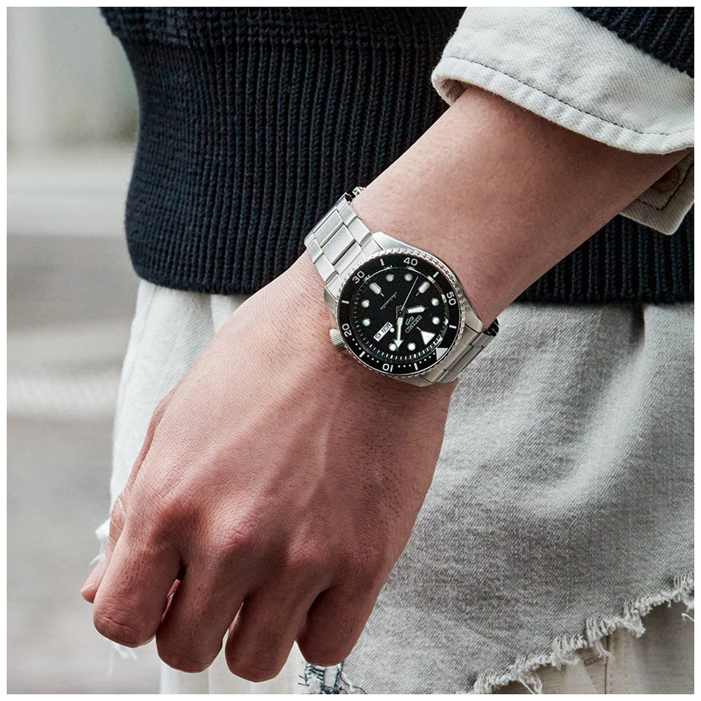 Ze dragen een Seiko 5 Sports herenhorloge SRPD55K1, een zilveren horloge met een zwarte wijzerplaat en witte markeringen van Seiko, en zijn gekleed in een zwarte trui met witte mouwen, en houden hun hand aan hun zijde. De roestvrijstalen kast glanst tegen de wazige, neutraal getinte achtergrond.