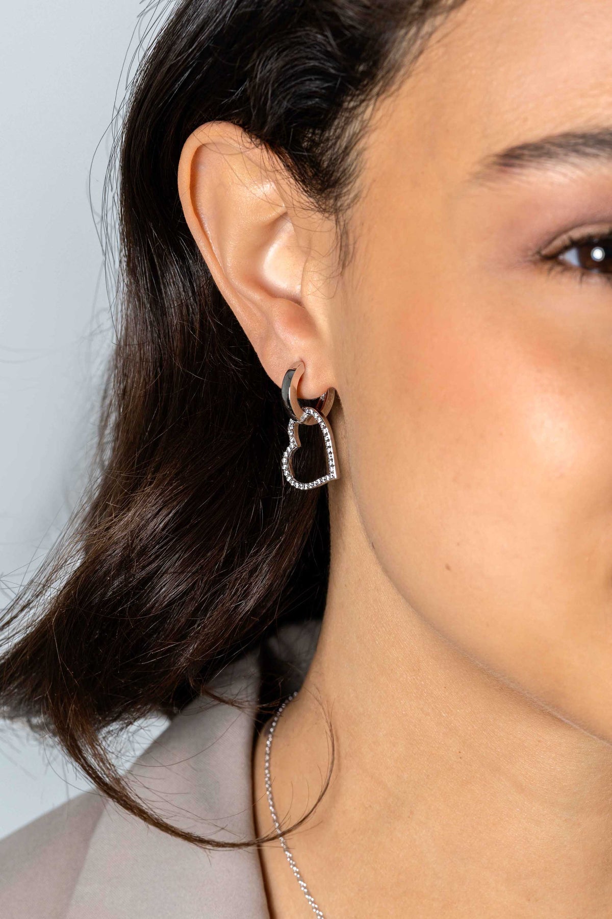 Close-up van het oor van een vrouw versierd met Zinzi Oorbedels zilver ZICH2447, een hartvormige zilveren oorbel met kleine edelsteentjes. Haar donkere, steile haar bedekt haar oor gedeeltelijk en ze draagt een fijne ketting.