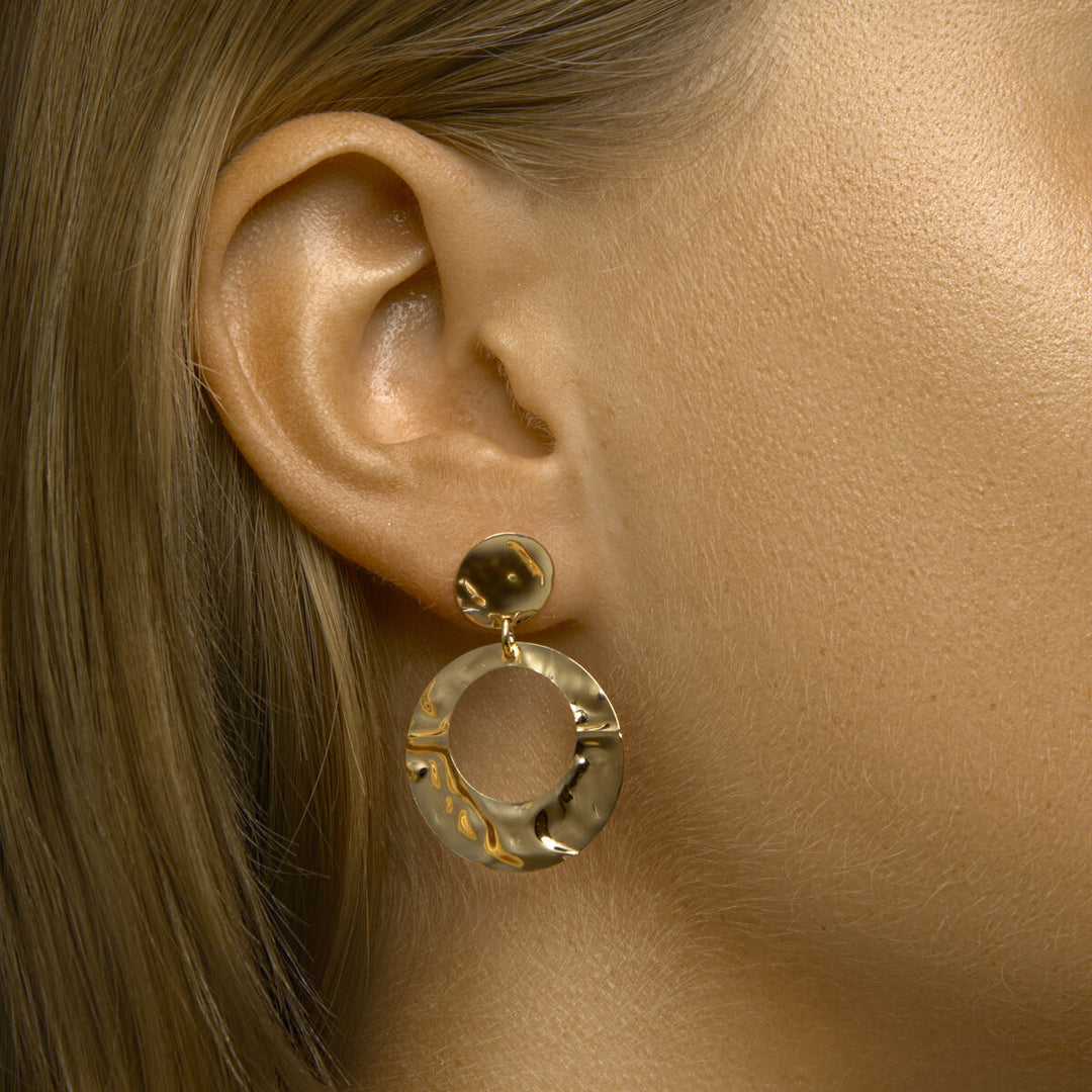 Close-up van het oor van een vrouw met Van der Kooij Collectie oorhangers gehamerd 1 micron zilver verguld (geel): gestructureerde, ronde oorbellen met een kleine ronde knop en een grotere open cirkel eronder. Haar haar en huid zijn lichtbruin.