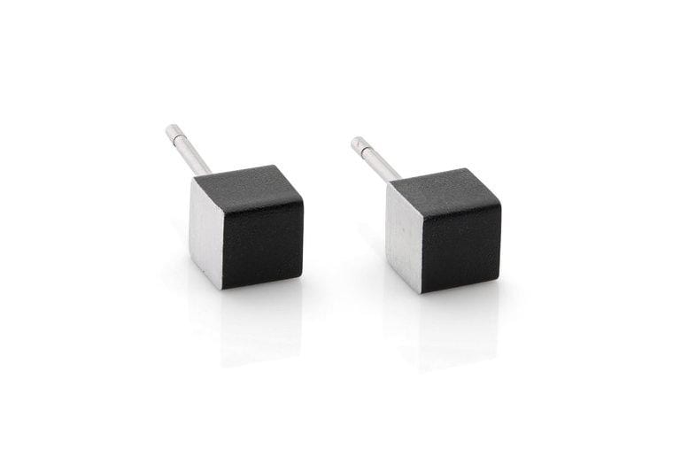 Twee kleine kubusvormige oorstekers met zwarte bovenkanten en zilveren steeltjes worden elegant tentoongesteld op een wit reflecterend oppervlak. De Oorsieraad Kubusjes Zwart O28Z van CLIC Creations zijn hypoallergeen en combineren comfort en stijl voor gevoelige oren.