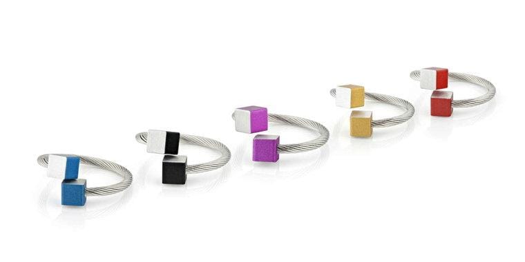 Vijf zilveren gedraaide draadringen van CLIC Creations zijn gemaakt van chirurgisch staal, elk met unieke ontwerpen met gekleurde kubussen - blauw, zwart, paars, goud en rood - aan de uiteinden. De Ring Kubusjes Blauw R4B toont deze tinten prachtig in een diagonale lijn tegen een witte achtergrond.