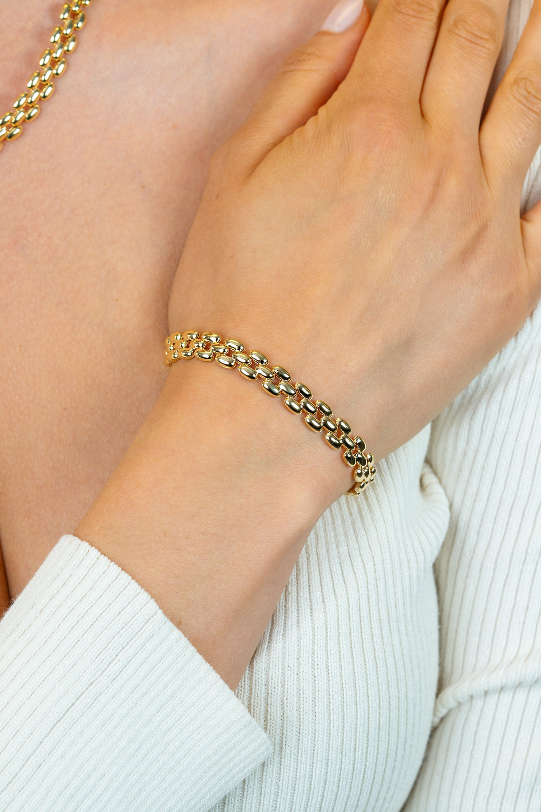 Een close-up van een persoon die de Zinzi Armband Gold Plated ZIA2764G en een bijpassende gouden ketting draagt, met één hand zachtjes rustend op de borst over een wit geribd kledingstuk.