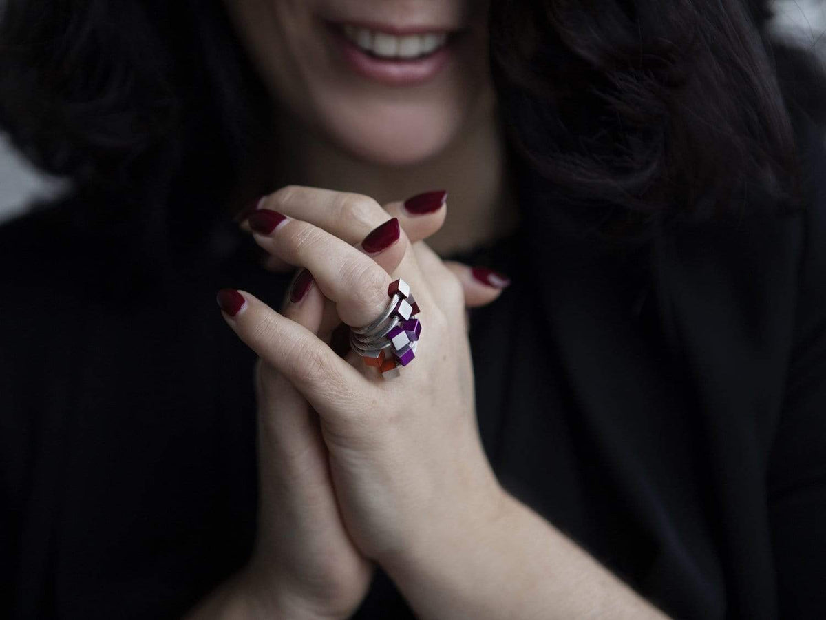 Een persoon met donker golvend haar lacht en vouwt zijn handen in elkaar. Ze dragen een uniek ontworpen Ring Kubusjes Blauw R4B van CLIC Creations, gemaakt van chirurgisch staal, naast kleurrijke ringen, terwijl donkerrode nagellak hun donkere outfit complementeert.