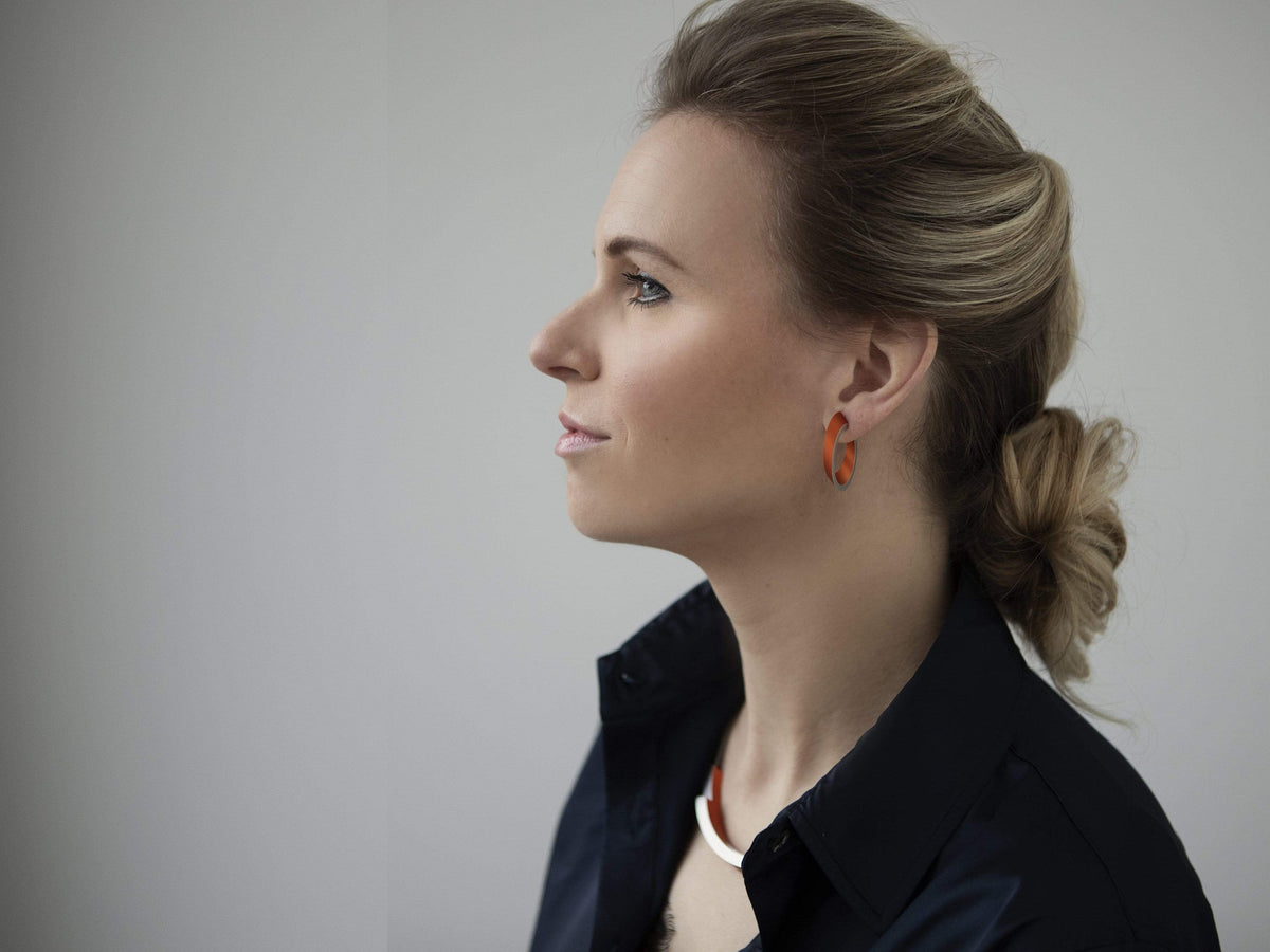 Een persoon met licht naar achteren gebonden haar, een zwart shirt en oranje oorbellen, staat tegen een effen achtergrond, aangevuld met een CLIC Creations Collier Bogen C74.
