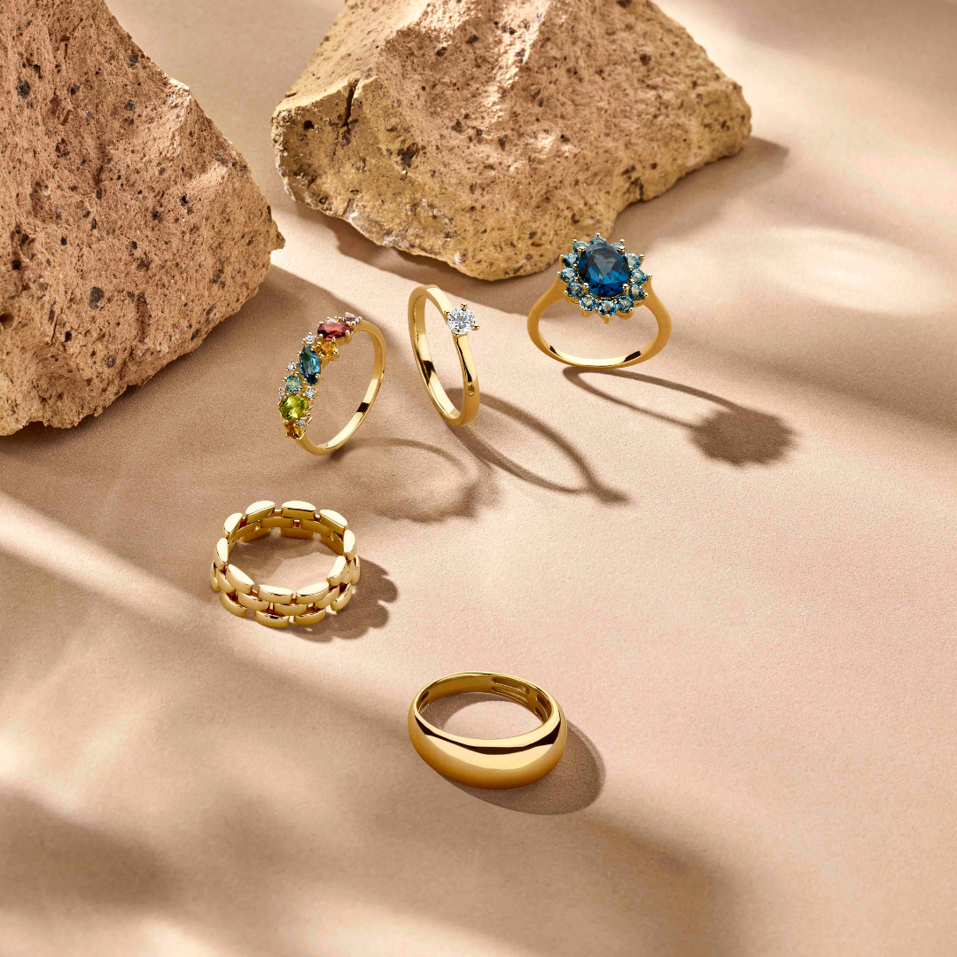 Vijf gouden ringen, waaronder de Van der Kooij Collectie ring met natuurlijke kleurstenen en 0,04ct diamant in 14K geelgoud, worden getoond op een beige oppervlak met twee beige rotsen met textuur die zachte schaduwen werpen op de achtergrond.