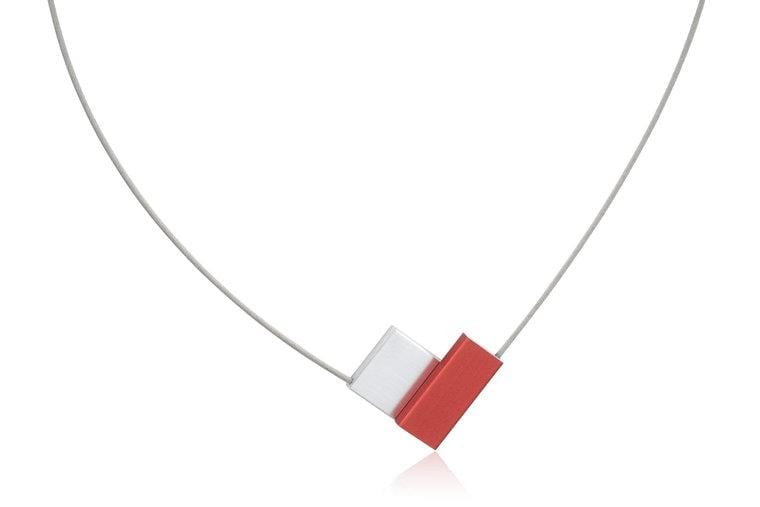 De Collier V-vorm Rood C150R van CLIC Creations is een minimalistische ketting met een uniek design met een dunne draad en twee aangrenzende kubussen als hanger, een zilveren en een rode, wat een moderne geometrische look creëert. Handgemaakt in Nederland, inclusief magnetische sluiting voor eenvoudig dragen.