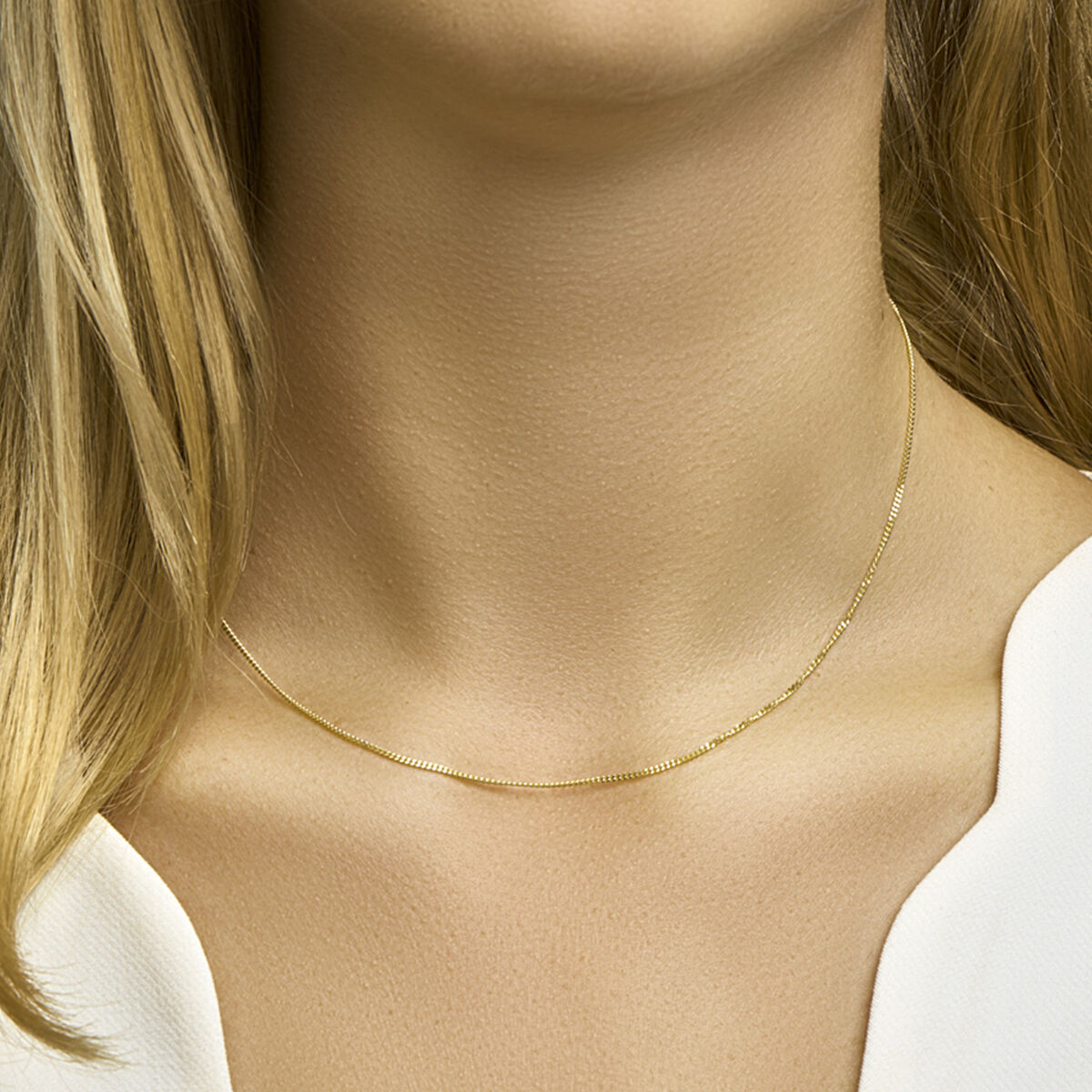 Een vrouw met lichtblond haar en een witte top draagt de Van der Kooij Collectie collier gourmette 4-zijdes geslepen 1,2 mm 14K geelgoud. De afbeelding benadrukt haar nek en borst, waardoor het subtiele gastronomische ontwerp goed naar voren komt.