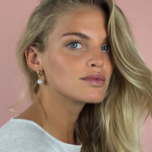 Een vrouw met lang blond haar in een wit topje draagt Zinzi Oorbellen Gold Plated Zich266 Wg oorbellen, poserend tegen een roze achtergrond.
