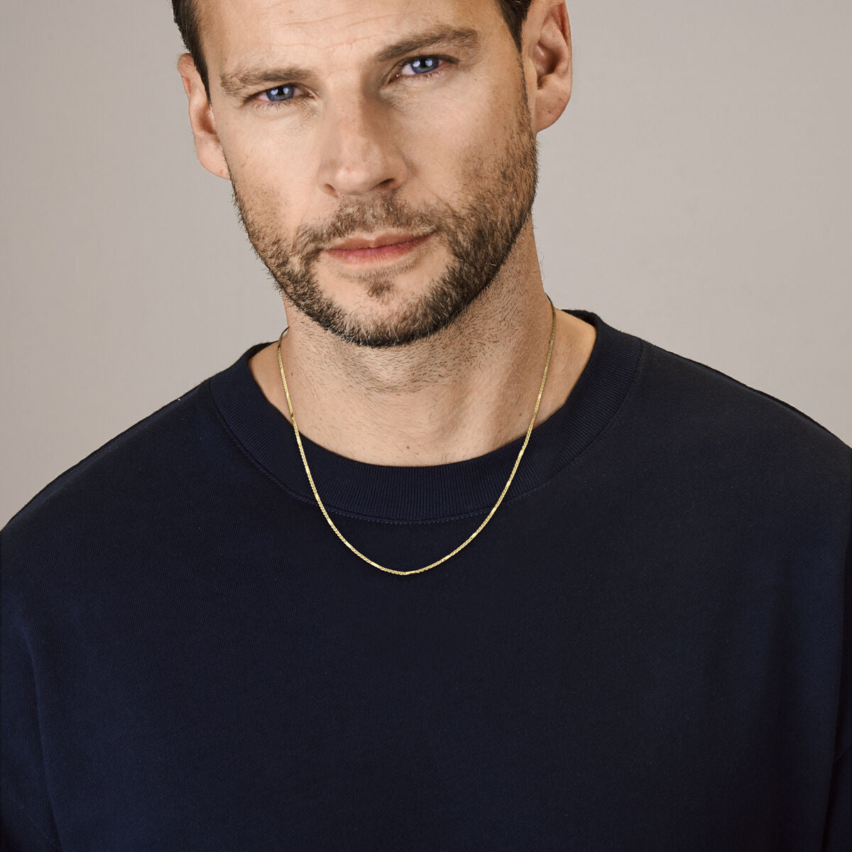 Een man met kort bruin haar, blauwe ogen en een getrimd baardje draagt een navy crewneck trui en een Van der Kooij Collectie 14K gouden ketting heren - collier valkenoog met tussenstuk 1,9 mm. Hij kijkt neutraal langs de camera tegen een effen achtergrond.
