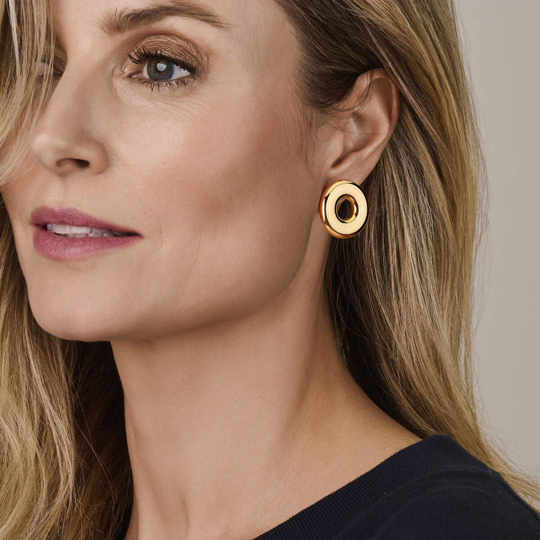 Een vrouw met een lichte huid en lang blond haar draagt een donker topje en Van der Kooij Collectie oorknoppen rondje 32 mm 1 micron zilver verguld (geel), grote ronde goudkleurige oorbellen met een zwart hart, die neutraal naar de zijkant kijken.