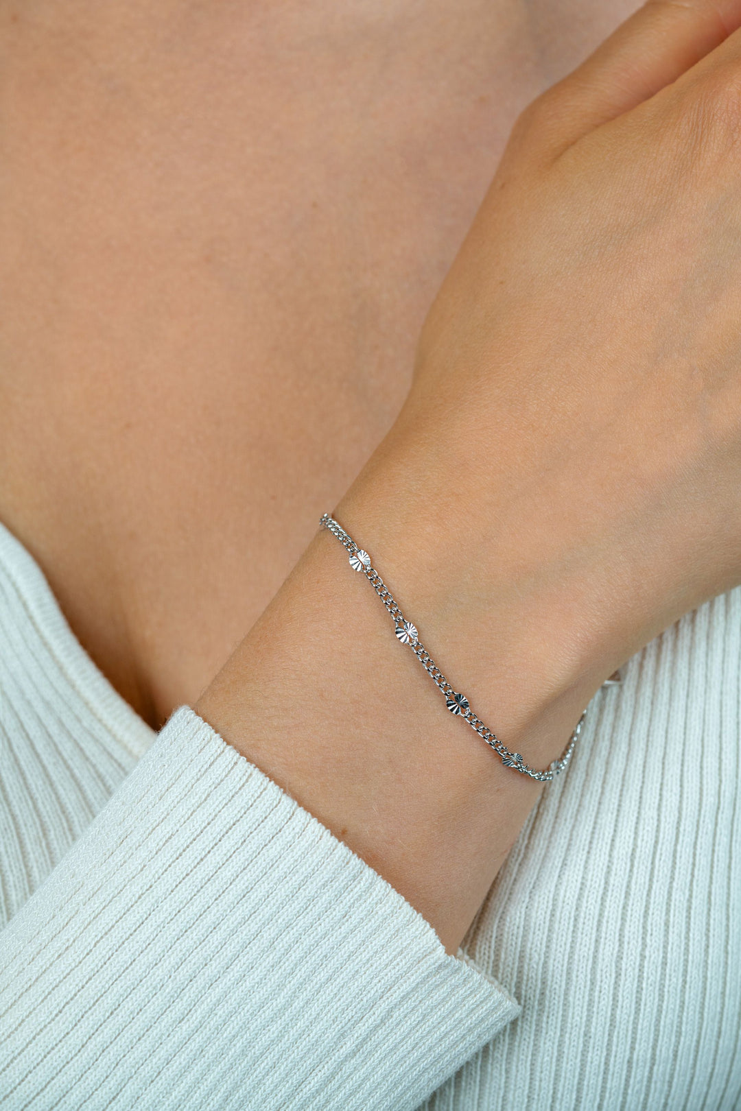 Een close-up van een pols die de Zinzi Armband zilver ZIA2713 van Zinzi draagt, een delicaat eerste gehalte zilveren gourmet schakelarmband met kleine bedeltjes en rodiumlaag, afgebeeld tegen een lichtbeige geribbelde top bij het sleutelbeen.