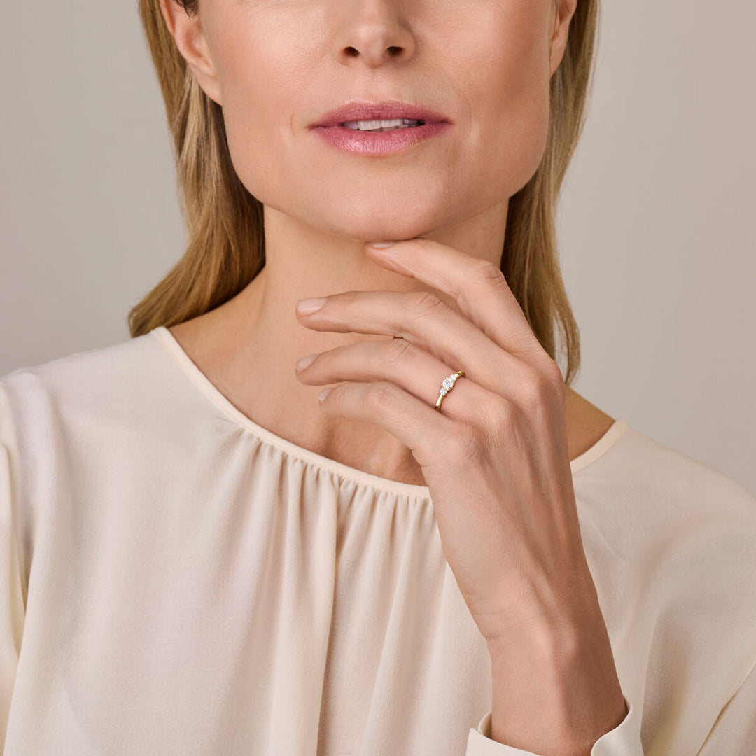 Een vrouw in een crèmekleurige blouse met lichtroze lippenstift raakt zachtjes haar kin aan en toont de Van der Kooij Collectie ring trilogie lab grown diamant 0.46ct H SI 14K geelgoud aan haar vinger, haar uitdrukking is kalm.
