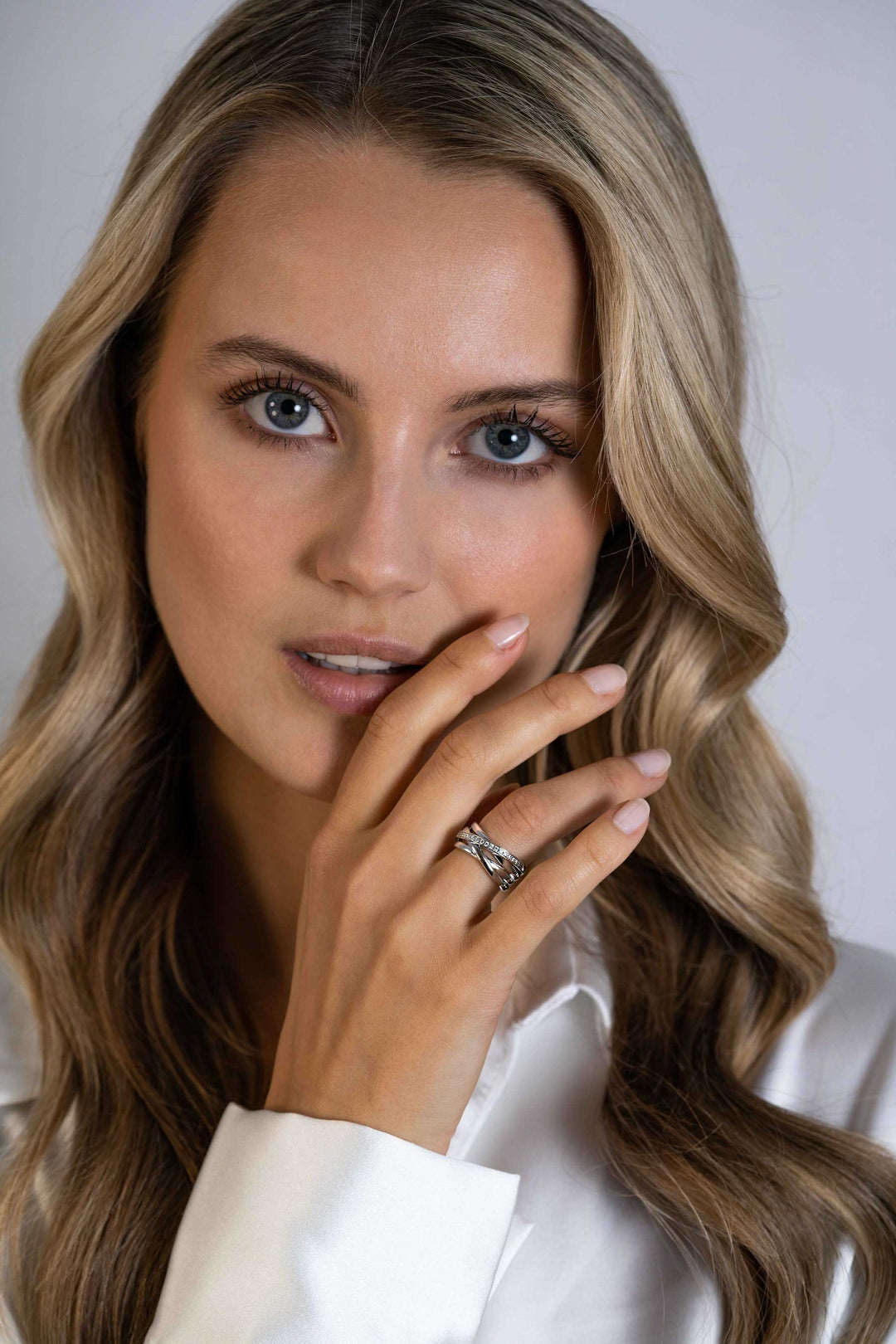 Een vrouw met golvend blond haar en blauwe ogen poseert met haar hand voor haar lippen en toont de Zinzi Ring zilver ZIR1790 van Zinzi aan haar vinger. Ze draagt een wit shirt en kijkt recht in de camera.
