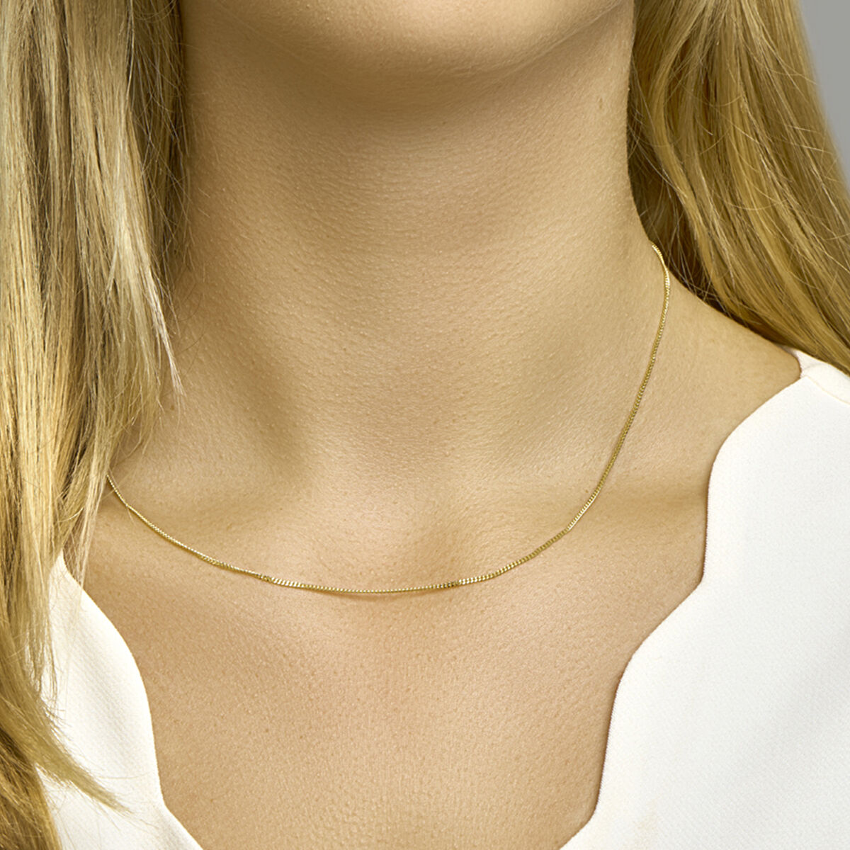 Een vrouw met lang blond haar draagt de Van der Kooij Collectie collier gourmette 4-zijden geslepen 1,2 mm in 14 karaat geelgoud om haar hals. Ze combineert het met een witte top met een subtiele V-hals.
