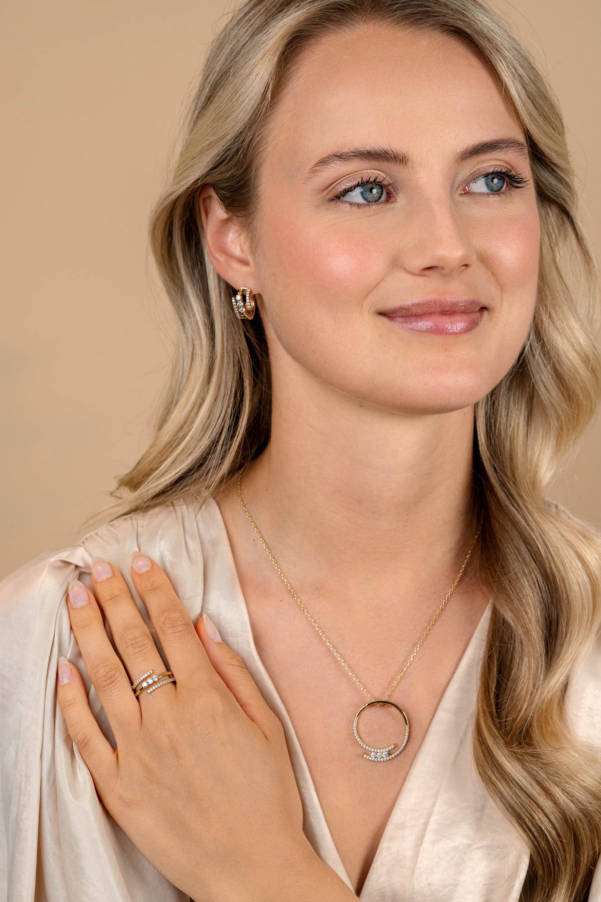 Een vrouw met lang blond haar draagt een lichtbeige satijnen blouse, gouden ringen en Zinzi oorringen zilver ZIO2645Y-verguld zilveren hoepeloorbellen van Zinzi-zacht glimlachend tegen een neutrale achtergrond.