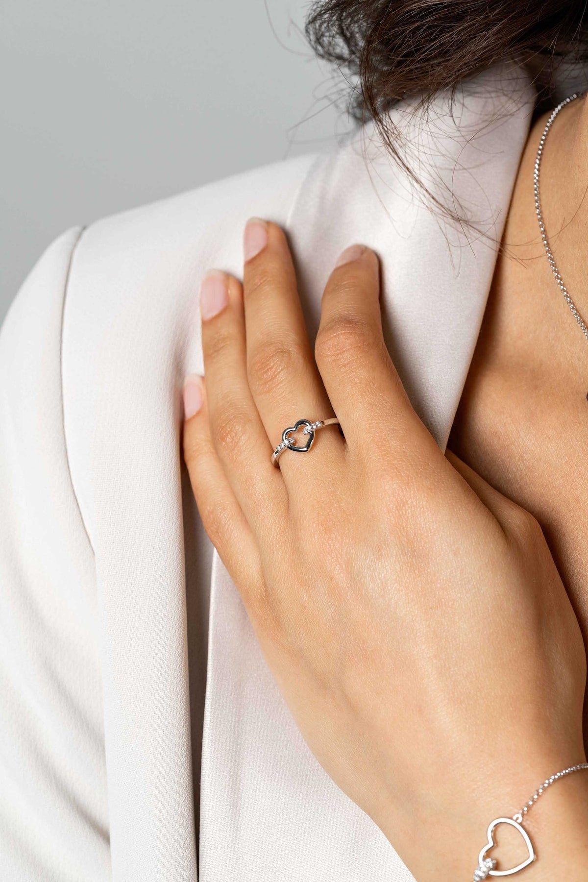 Een close-up van de hand van een vrouw die de Zinzi Ring Zilver Zir1755 draagt, met een open hart en witte zirkonia's, rustend op een witte blazer. Ze draagt ook een hartjesarmband en een delicate halsketting, met natuurlijke nagels en zachte belichting.