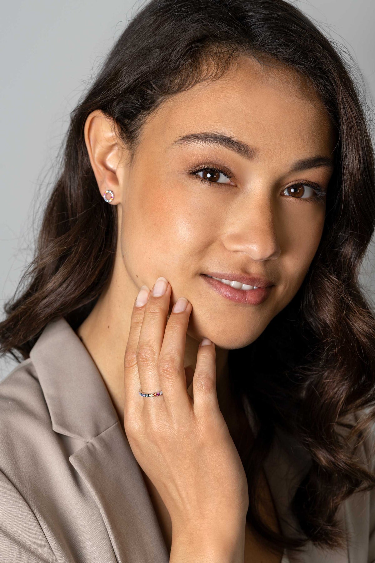 Een vrouw met lang, golvend bruin haar glimlacht zachtjes. Ze draagt een taupe blazer, kleine oorbellen en de Zinzi Ring Zilver Zir2486 van Zinzi, haar hand rust op haar kin tegen een lichtgrijze achtergrond.
