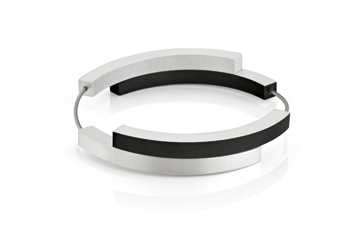 De Armband Vier bogen Zwart A32Z van CLIC Creations is een moderne, minimalistische bangle met afwisselende segmenten van zilver en zwart, verbonden door een dunne draad. Het vormt een halfopen cirkel voor een strakke, stijlvolle look. Handgemaakt in Nederland, het wordt weergegeven op een wit reflecterend oppervlak.
