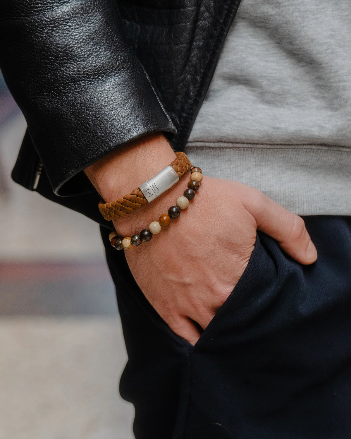 Een persoon draagt een zwart leren jasje en een grijze trui en heeft zijn hand in zijn zak. Hij accentueert zijn look op elegante wijze met een AZE Jewels Olympus Kralen armband voor heren en een bruine gevlochten armband om de pols.