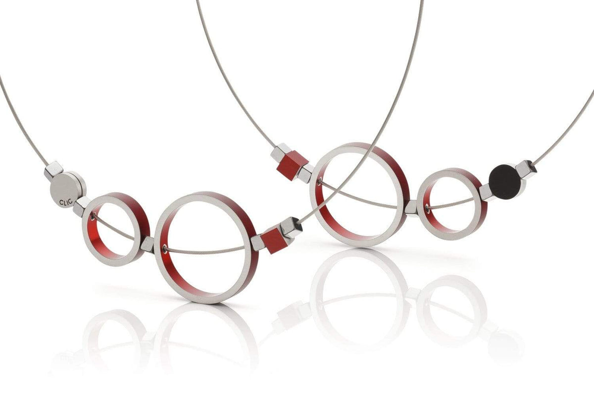 De Collier Ringen Rood C186R van CLIC Creations heeft een minimalistisch ontwerp met onderling verbonden cirkels en vierkanten. Metalen elementen hebben rode accenten en zijn verbonden op dunne draadkettingen met een magnetische sluiting, allemaal tegen een overzichtelijke witte achtergrond voor een moderne artistieke flair.