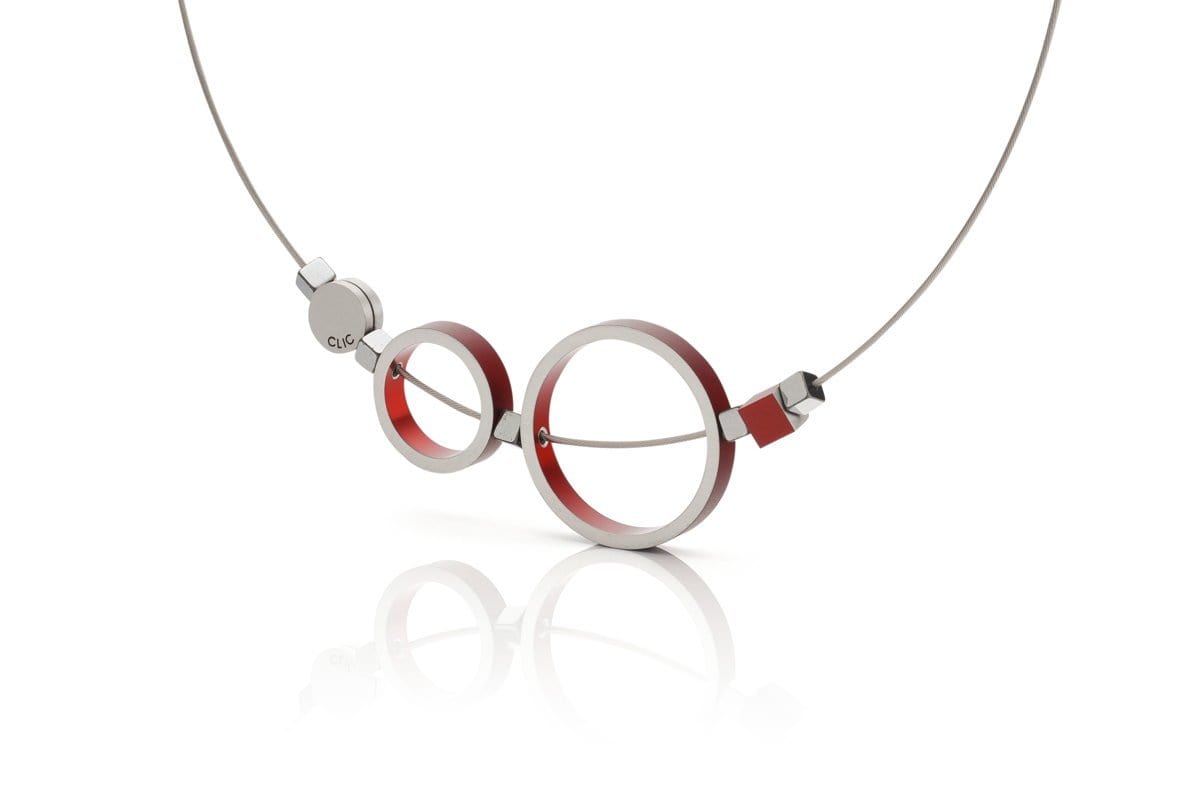 De Collier Ringen Rood C186R van CLIC Creations is een moderne ketting met een uniek ontwerp van twee metalen cirkels met rode accenten die lijken op een bril, hangend aan een dunne draadketting. Een subtiel logo siert de kleine ronde bevestiging aan de linkerkant, die allemaal prachtig reflecteren op een wit oppervlak.