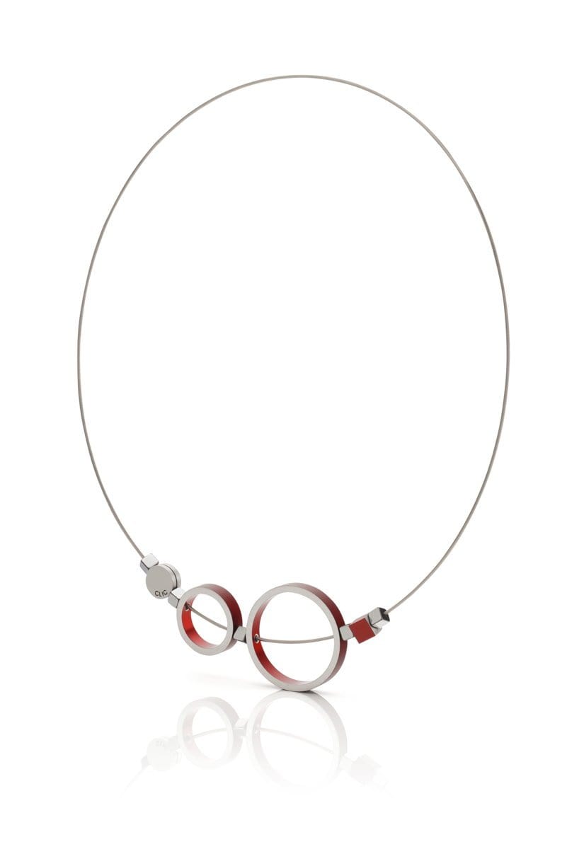 De Collier Ringen Rood C186R van CLIC Creations is een minimalistische ketting met twee grote, in elkaar grijpende, zilveren ronde hangers met rode accenten. Deze hangen aan een dunne, zilveren ketting met een magnetische sluiting aan de zijkant en vormen een witte achtergrond.