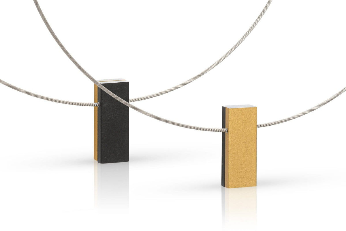 De Collier twee gekleurde rechthoeken C232GZ van CLIC Creations heeft zwarte en gele rechthoekige blokken die met elkaar verbonden zijn door dunne, gebogen metalen draden met een magnetische sluiting. Handgemaakt in Nederland, vormt het strakke, minimalistische lijnen op een reflecterend oppervlak.