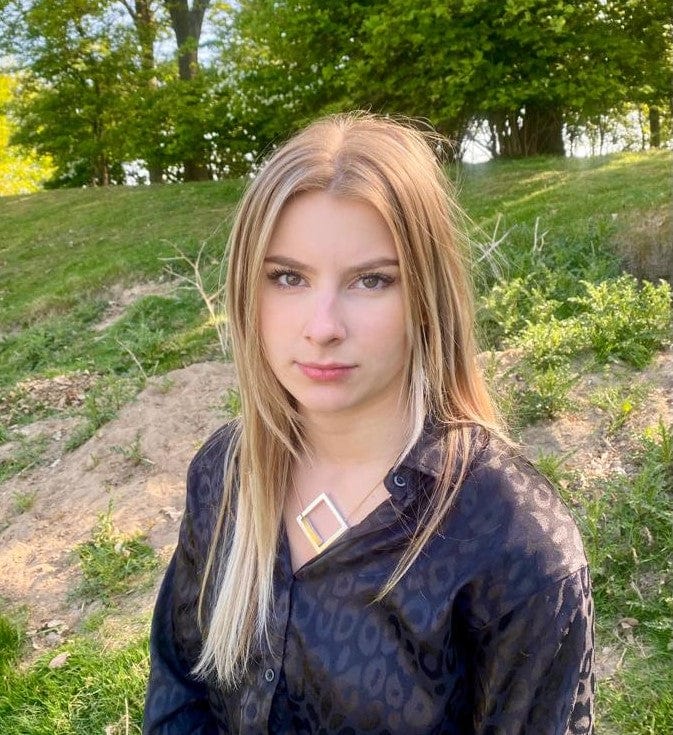 Een jonge vrouw met lang blond haar zit op een grasheuvel, draagt een donker gebloemde blouse en de Collier Vierkant met boogje Goud|Geel C252G van CLIC Creations. Haar ketting is handgemaakt in Nederland en heeft een elegante magnetische sluiting. Bomen en groen creëren een serene achtergrond.
