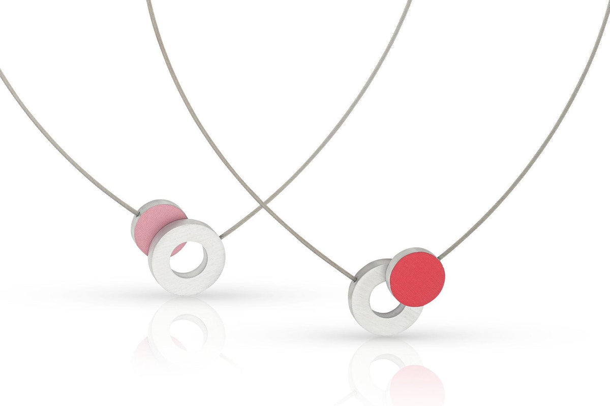 De Collier Gekleurd rondje en een ring C265R van CLIC Creations bestaat uit twee minimalistische kettingen met dunne zilveren kettingen en ronde hangers met een roze en wit geometrisch ontwerp. Deze unieke stukken zijn handgemaakt in Nederland en rusten op een witte achtergrond voor een moderne, stijlvolle look.
