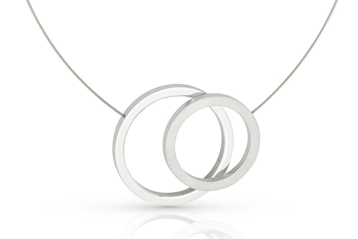 De CLIC Creations Collier grote ringen C267 heeft een modern design met twee in elkaar grijpende zilveren cirkels op een dunne ketting en een magnetische sluiting. De gladde, gepolijste ringen bieden elegantie tegen een effen witte achtergrond.